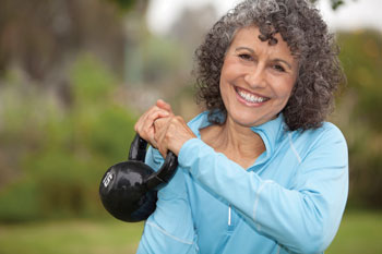 An older woman outdoors wearing a sweatshirt, holding a kettlebell weight up with her two hands.